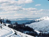 San Carlos de Bariloche cubierta de nieve vista desde la altura de una montaña, al fondo el Lago Nahuel Huapi y la Cordillera de los Andes